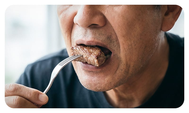 A happy elderly man confidently chewing and eating meat, representing restored chewing function after dental implant treatment.