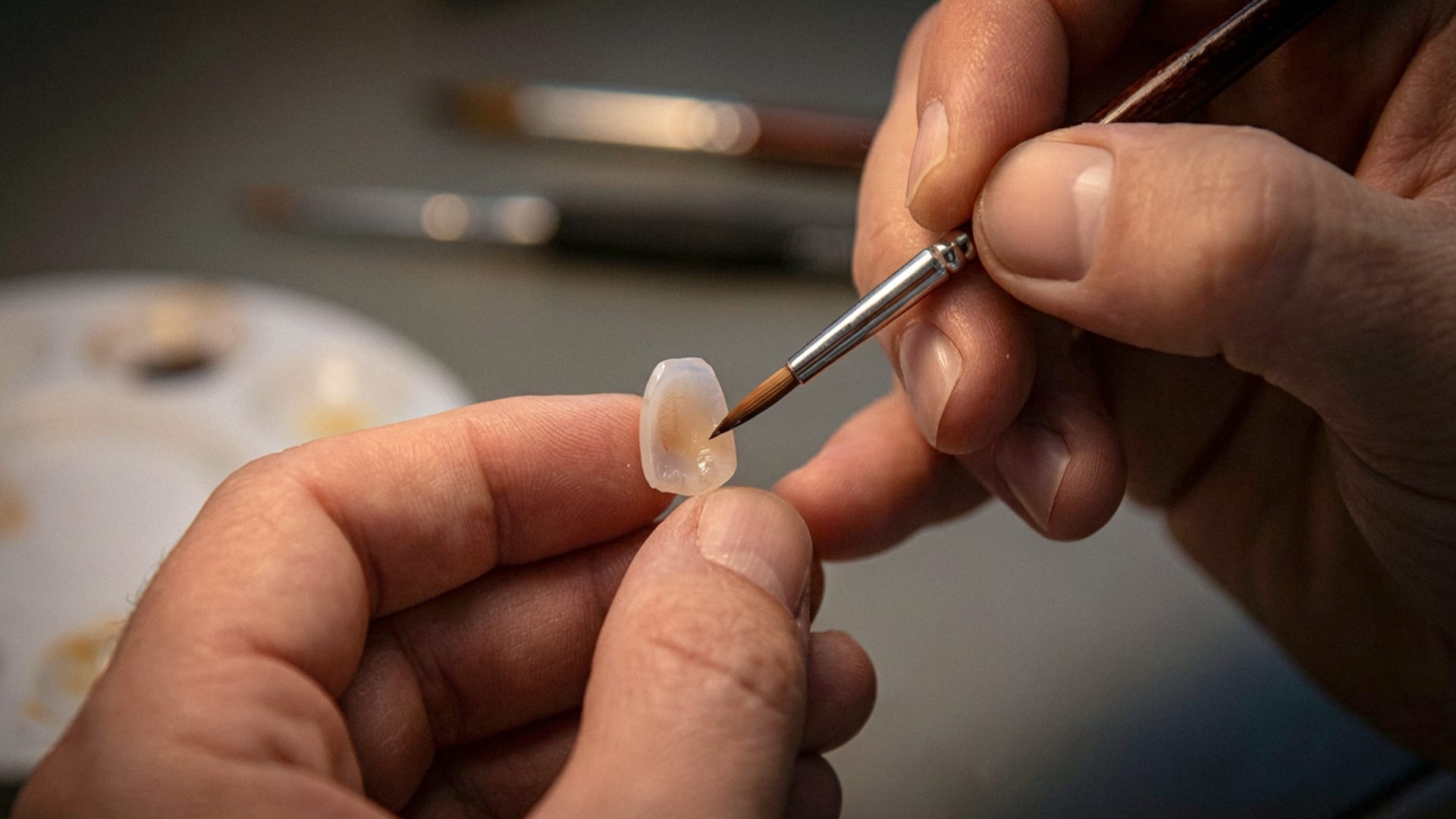 A close-up shot of a dental technician's hands delicately finishing and staining a small laminate restoration using a fine dental brush.
