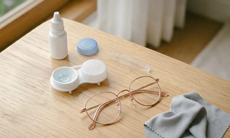 An array of eye care products including eye drop bottles, a contact lens case, lenses, tweezers, and glasses